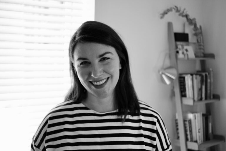 Laura Stannard smiling in a black and white photo, wearing a striped top, with a ladder bookshelf behind her