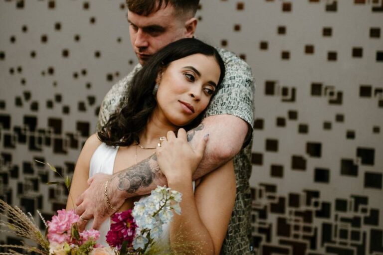 A man hugs a woman from behind as she holds a bouquet of pink and blue flowers, in front of a patterned wall