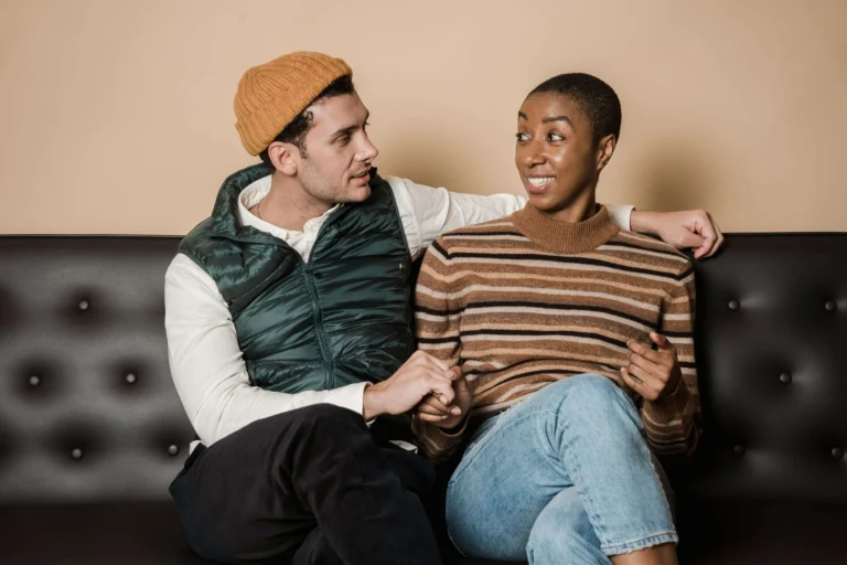 A couple holding hands on a sofa, the man in a beanie and gilet and the woman smiling at him