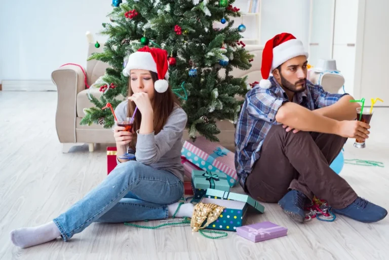 A couple in Santa hats sitting back to back on the floor by a Christmas tree, looking away from each other
