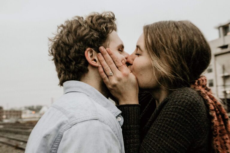 A woman with her hand on a man's cheek, kissing him outdoors at dusk