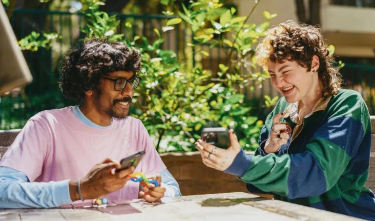 Two friends laughing at their phones at an outdoor table, one holding a rainbow fidget toy
